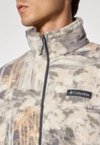 Fleecejakke i beige med grå og taupe mønstre, glidelås i kragen, og en svart Columbia-logo på brystet. Myk tekstur og avslappet design.
