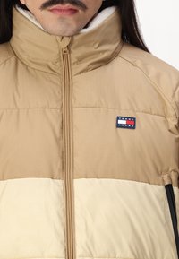 Tan and cream puffer jacket with a high collar, zip closure, and stitched details. Features a black accent and a logo patch on the chest.