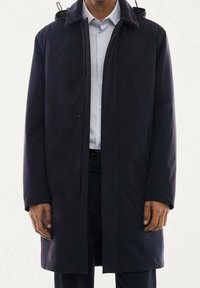 Abrigo azul marino con capucha, con corte recto, cierre de botones de presión y mangas largas. Fabricado con una tela lisa, se lleva sobre una camisa ligera.