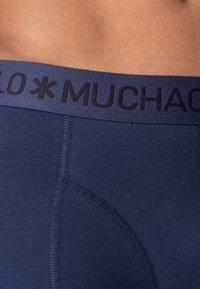 Dark blue boxer briefs with a black elastic waistband featuring the text "MUCHACHO." The fabric is smooth and stretchy, showing stitched seams.