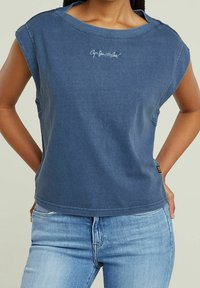 Žena v bezramenném, vybledlém modrém tričku s kulatým výstřihem a malým logem na hrudi, v kombinaci s světle modrými džíny, s rukama položenýma na bocích.