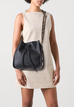 Borsa a secchiello in pelle nera con chiusura a coulisse, dotata di una tracolla con logo e un design strutturato. Abbinata a un vestito a quadretti leggeri.