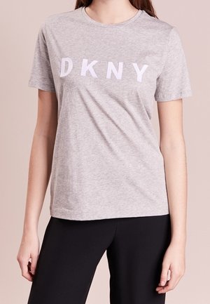Person trägt ein hellgraues Kurzarm-T-Shirt mit weißen "DKNY" Buchstaben und schwarze Hose, steht vor einfachem Hintergrund.
