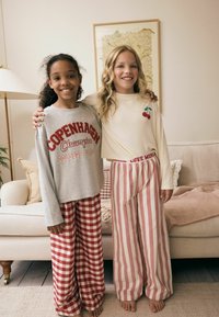 Zwei Kinder tragen Schlafanzüge. Das eine Kind hat ein graues Langarmshirt mit rotem Schriftzug und karierter rot-weißer Hose an. Das andere trägt ein cremefarbenes Oberteil mit einem Kirschdesign und rot-gestreifte Hosen.