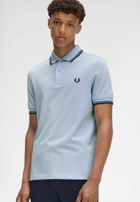 Hellblaues Polohemd aus Baumwolle, mit einem schwarzen Logo, einem schwarz-navy gestreiften Kragen und kurzen Ärmeln. Glatte Textur und klassischer Schnitt.