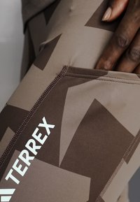 Gros plan d'une main posée sur un tissu marron et beige à motif géométrique avec le mot « TERREX » imprimé en blanc.