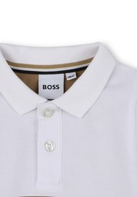 Witte polo met een gestructureerde weefsel, voorzien van een bruin en zwart gestreepte kraag en een enkele knoopsluiting. Merkenlabel zichtbaar.