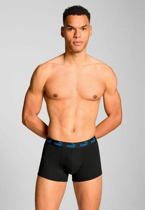 Black boxer briefs with a blue Puma logo waistband, fitted design, soft texture, and mid-thigh length. Model standing with hands behind back.