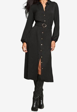 Robe noire boutonnée avec manches longues, taille ceinturée avec une boucle circulaire, fente haute jusqu'à la cuisse, et accents de boutons dorés.