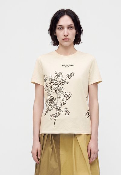 Jeune femme aux cheveux courts et foncés portant un t-shirt beige à motif floral et une jupe plissée couleur moutarde, debout devant un fond clair uni.