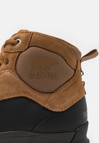 Czekoladowe zamszowe, wodoodporne buty z czarną gumową podeszwą. Posiadają teksturowaną, tanową wstawkę z logo niedźwiedzia oraz wzmocnione wsparcie kostki.