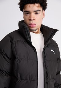 Chaqueta de pluma negra con cierre de cremallera, cuello alto y detalle de logo blanco. Tejido suave y texturizado con paneles cosidos para aislamiento.