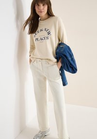 Femme portant un sweat-shirt beige avec le texte « Seek New Places », un pantalon crème, des baskets blanches, tenant une veste en jean bleue pliée, debout contre un mur.