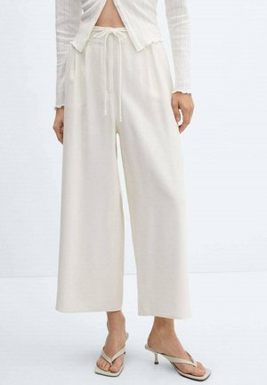 Femme portant un pantalon ample crème à jambes larges avec une taille à cordon et des sandales crème à talons en forme de tongs, debout devant un fond clair uni.