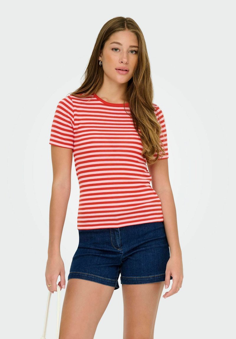 Jonge vrouw met lang haar, gekleed in een rood-wit gestreept T-shirt en donkere denim shorts, die een wit tashandvat vasthoudt.