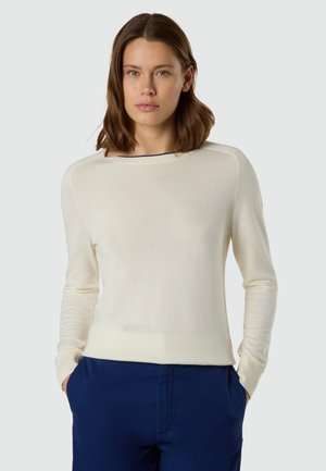 Donna con capelli castani fino alle spalle, che indossa un maglione crema a maniche lunghe e pantaloni blu navy, in piedi con le mani nelle tasche.