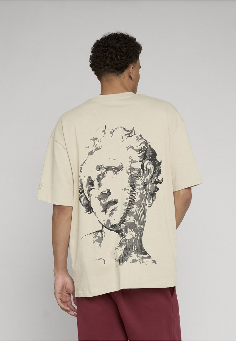 Homme portant un t-shirt beige oversize avec un grand croquis noir et gris d’un buste de sculpture classique dans le dos, associé à un short rouge.