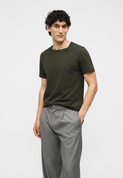 Νέος άνδρας με κοντά μαύρα σγουρά μαλλιά, που φοράει σκούρο πράσινο μπλουζάκι και ανοιχτογκρι πιετέ παντελόνια, στέκεται με το ένα χέρι στην τσέπη.