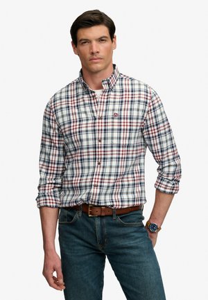 Hombre con camisa de cuadros en blanco, rojo y azul abotonada, con las mangas remangadas, vaqueros azules, cinturón marrón y reloj de pulsera sobre un fondo liso.