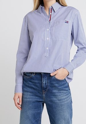 Femme portant une chemise boutonnée à rayures verticales bleues et blanches avec poche poitrine et jean bleu, main dans la poche.