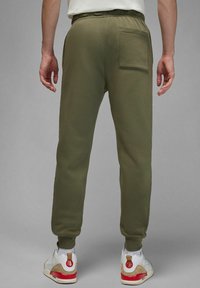 Persona che indossa pantaloni jogger verde oliva con tasca posteriore e sneakers bianche con dettagli rossi e beige, in piedi contro uno sfondo grigio.