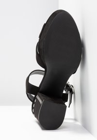 Sandale en daim noir avec talon bloc, clous argentés sur le côté du talon et bride de cheville avec boucle, vue de la semelle sur fond blanc.
