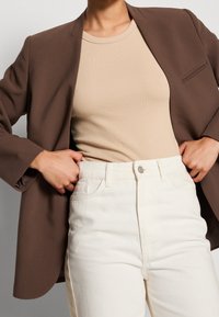 Blazer marrón, top beige de canalé y jeans blancos de talle alto. El blazer tiene un solo bolsillo; tela lisa; ajuste casual y entallado.