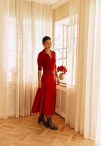 Frau in einem roten Kleid mit schwarzem Gürtel und Knöchelstiefeln steht neben durchsichtigen Vorhängen und einer Fensterbank mit einem Blumentopf und Zweigen.