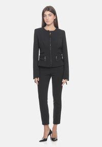 Giacca nera aderente con colletto rotondo, chiusura con zip e due tasche frontali. Abbinata a pantaloni neri slim e décolleté neri a punta.
