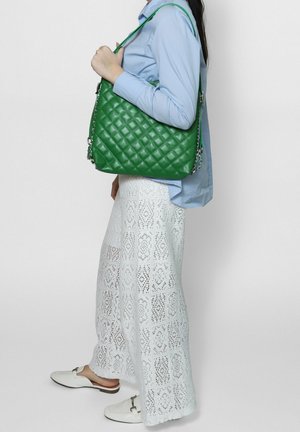 Donna con camicia blu e gonna di pizzo bianco che porta una grande borsa trapuntata verde sulla spalla.