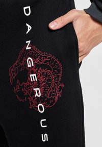 Schwarze Jogginghosen mit einem roten Drachenmotiv und dem Wort "DANGEROUS" in Weiß an der Seite, aus einem gerippten Material gefertigt.