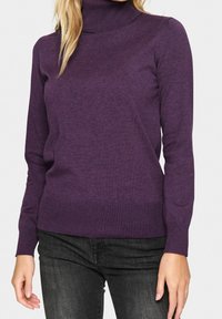 Femme portant un pull à col roulé violet ajusté avec des poignets et un ourlet côtelés, associé à un jean noir délavé, debout devant un fond uni.