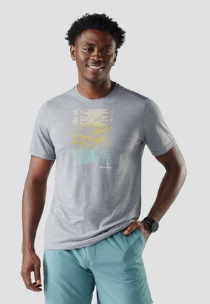 Uomo sorridente che indossa una t-shirt grafica grigio chiaro e pantaloncini turchese, con una mano in tasca e un orologio da polso nero al polso sinistro.