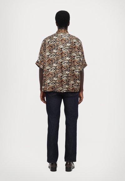 Košile s krátkým rukávem v černé barvě s vířivým vzorem v oranžové a krémové barvě; v kombinaci s tmavě modrými džíny; pohled zezadu.