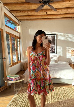 Frau in einem farbenfrohen gemusterten Kleid macht ein Spiegelselfie in einem hellen, rustikalen Schlafzimmer mit Holzdecke und großen Fenstern.