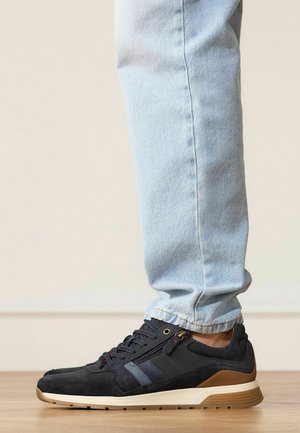 Persoon draagt lichtblauwe spijkerbroek en zwarte sneakers met een beige en witte zool, staand op een houten vloer tegen een effen beige muur.
