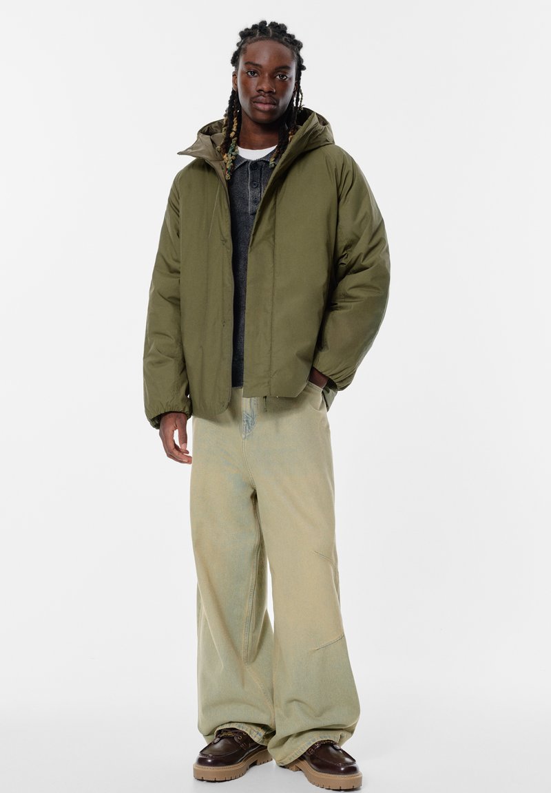 Giacca a vento verde oliva con cappuccio, abbinata a pantaloni larghi giallo chiaro e scarpe marrone scuro. Maglia basic grigia sotto.