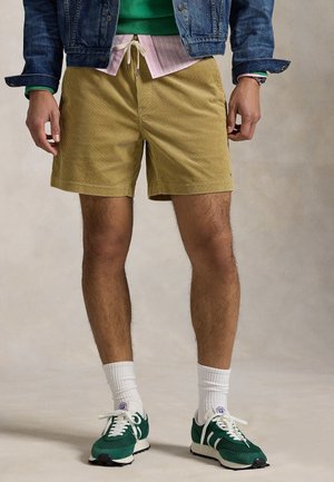 Mand iført beige manchester-shorts, grønne sneakers med hvide snørebånd, hvide sokker, stribet skjorte, grøn sweater og denimjakke.