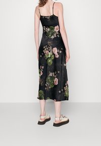 Kvinnor som bär en svart midiklänning med gröna och rosa blommönster och vita platta sandaler, stående mot en enfärgad ljusgrå bakgrund.