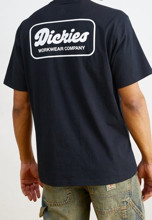 Mężczyzna w czarnej koszulce Dickies z logo i napisem "Workwear Company" na plecach, w połączeniu z zielonkawymi, zużytymi szortami dżinsowymi.