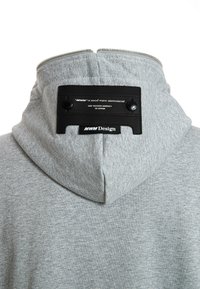 Sudadera gris con un tejido texturizado, que presenta una etiqueta de cuero negra en la parte posterior con el logo y botones a presión. Cremallera en el cuello para un ajuste ajustable.