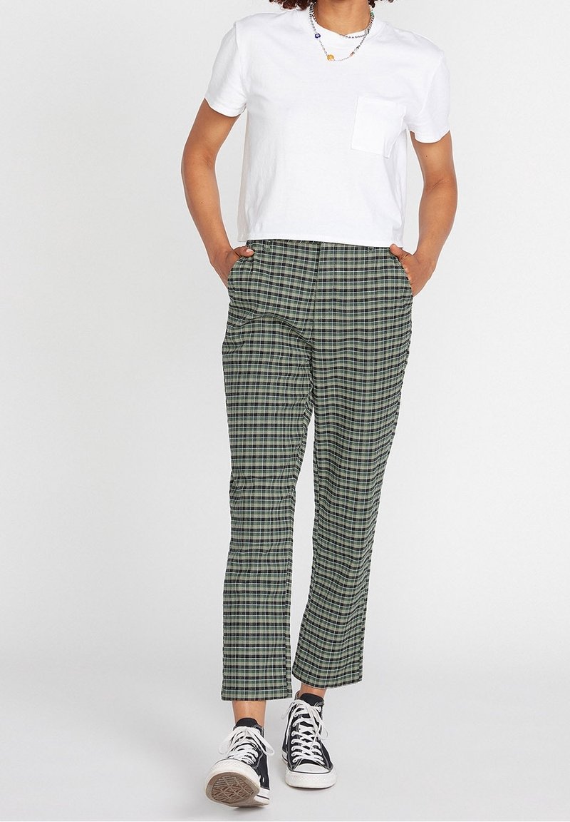 Personne portant une chemise blanche à manches courtes, un pantalon à carreaux verts et noirs, des baskets hautes noires et des colliers de perles superposés, debout les mains dans les poches.