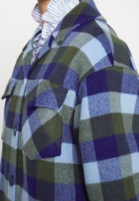Uldternet skjortejakke i nuancer af blå, grøn og lilla. Har store klaplommer, knaplukning foran og spids krave. Tekstureret stof.