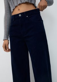 Pantalons larges en velours côtelé bleu marine avec une surface texturée, taille haute, fermeture à bouton et poches avant ; portés avec un pull cropped gris.