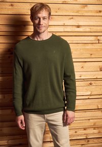 Pull en maille côtelée vert foncé avec des manches longues et un col rond, associé à un pantalon beige clair devant un fond en bois.