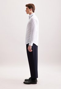 Uomo in piedi di lato che indossa una camicia da uomo azzurro chiaro, pantaloni scuri su misura e scarpe eleganti in pelle nere, su uno sfondo neutro.