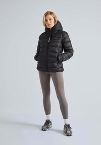 Svart puffjacka med huva, dragkedjestängning och logodetalj. Har en quiltad design och sidofickor, tillsammans med ljusbruna leggings och sneakers.