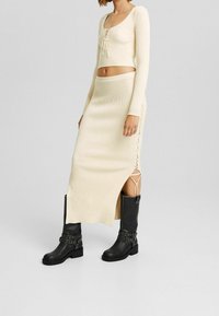 Kobieta w kremowym, prążkowanym topie w stylu crop z dopasowaną spódnicą z bocznymi sznurowaniami, w połączeniu z czarnymi, sięgającymi do połowy łydki, skórzanymi botkami na klamry.