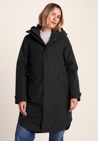 Woman wearing a long black hooded winter coat over a light purple sweater and blue jeans, standing against a neutral background.