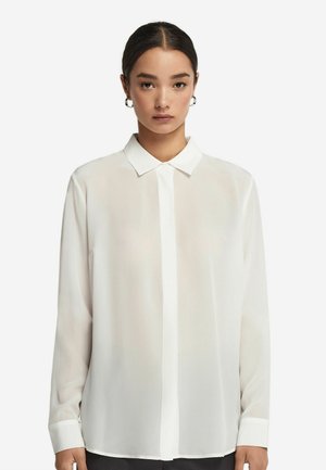 Młoda kobieta z ciemnymi włosami w niskim kokie nosząca srebrne kolczyki w kształcie obręczy i długą, białą bluzkę z kołnierzykiem na białym tle.